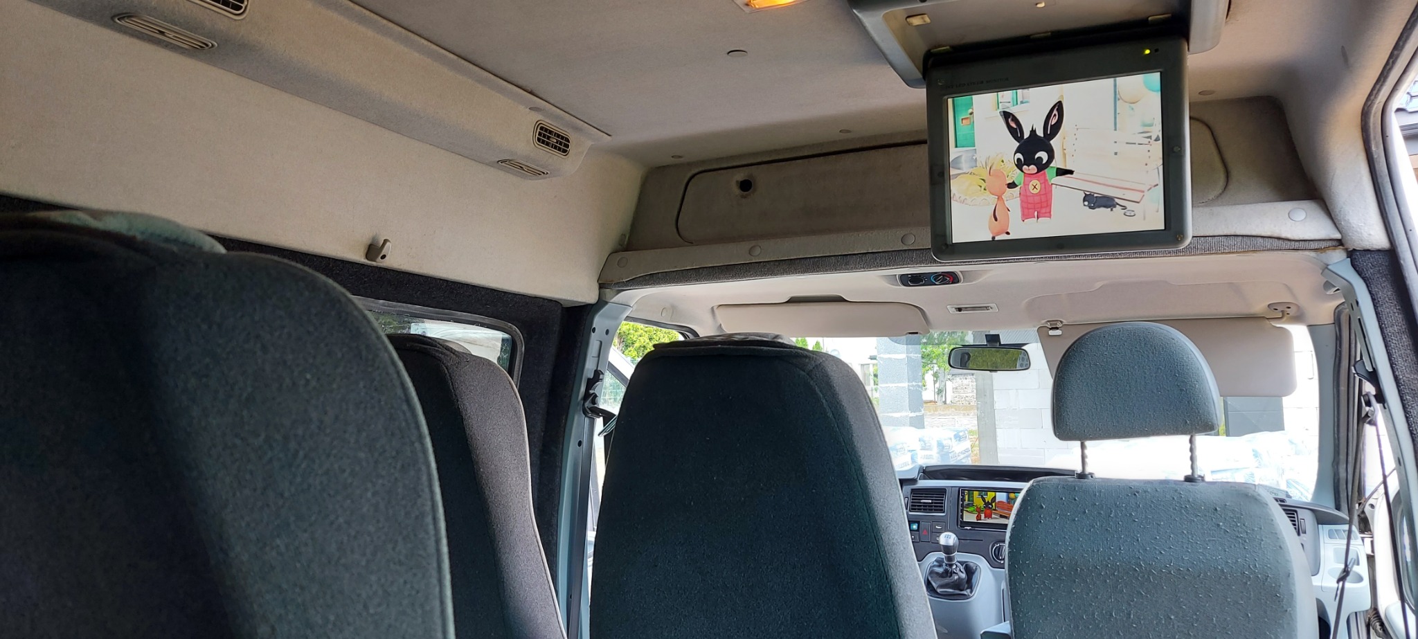 Wnętrze busa z zamontowanym monitorem sufitowym wyświetlającym bajkę dla dzieci, widok z tylnych siedzeń na deskę rozdzielczą i przednią szybę.