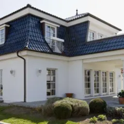 Biały dom z ciemnym, błyszczącym dachem z lukarnami i ozdobnym balkonem. Widoczne fragmenty elewacji, okna i zagospodarowanie terenu.