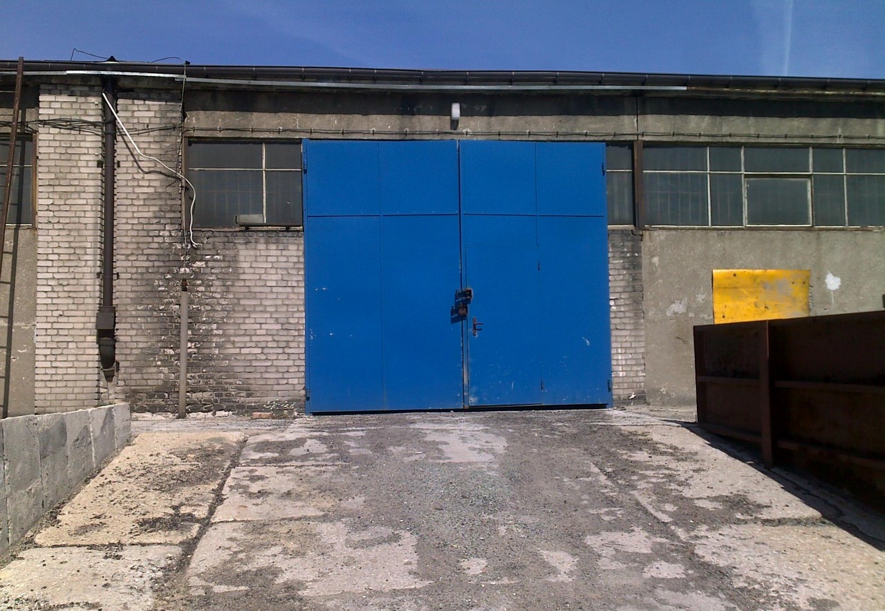 Duże, niebieskie, dwuskrzydłowe wrota magazynowe z zamkiem na tle ceglano-betonowego budynku z oknami, widoczne ślady wilgoci na murze i rampa wjazdowa.