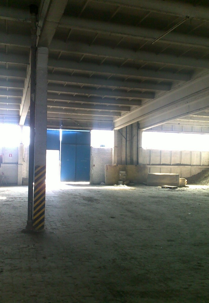 Wnętrze hali magazynowej z betonową konstrukcją stropu, widoczne niebieskie dwuskrzydłowe drzwi w oddali i słup oznaczony żółto-czarnymi pasami.