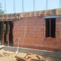 Surowy mur z czerwonej cegły ceramicznej, z drewnianym szalunkiem i podporami w otworach okiennych i drzwiowych, widoczne liny dźwigu nad budynkiem.