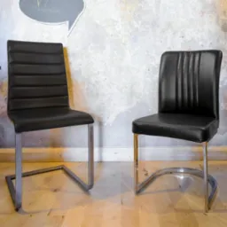 Dwa czarne krzesła z metalowymi nogami o różnych kształtach, ustawione na drewnianej podłodze na tle ściany z efektem spękań i elementem graficznym dymka.