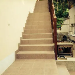 Wyłożone jasnymi, kwadratowymi płytkami schody zewnętrzne z drewnianą barierką. Na podeście widoczna packa do fugowania. Tło stanowią drzewa i fragment budynku.