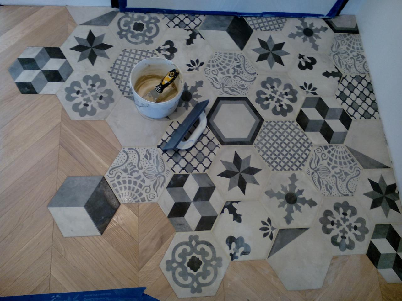 Układanie heksagonalnych płytek podłogowych z różnymi wzorami w łazience, widoczny kubeł z klejem i szpachelka, fragment drewnianej podłogi ułożonej w jodełkę.