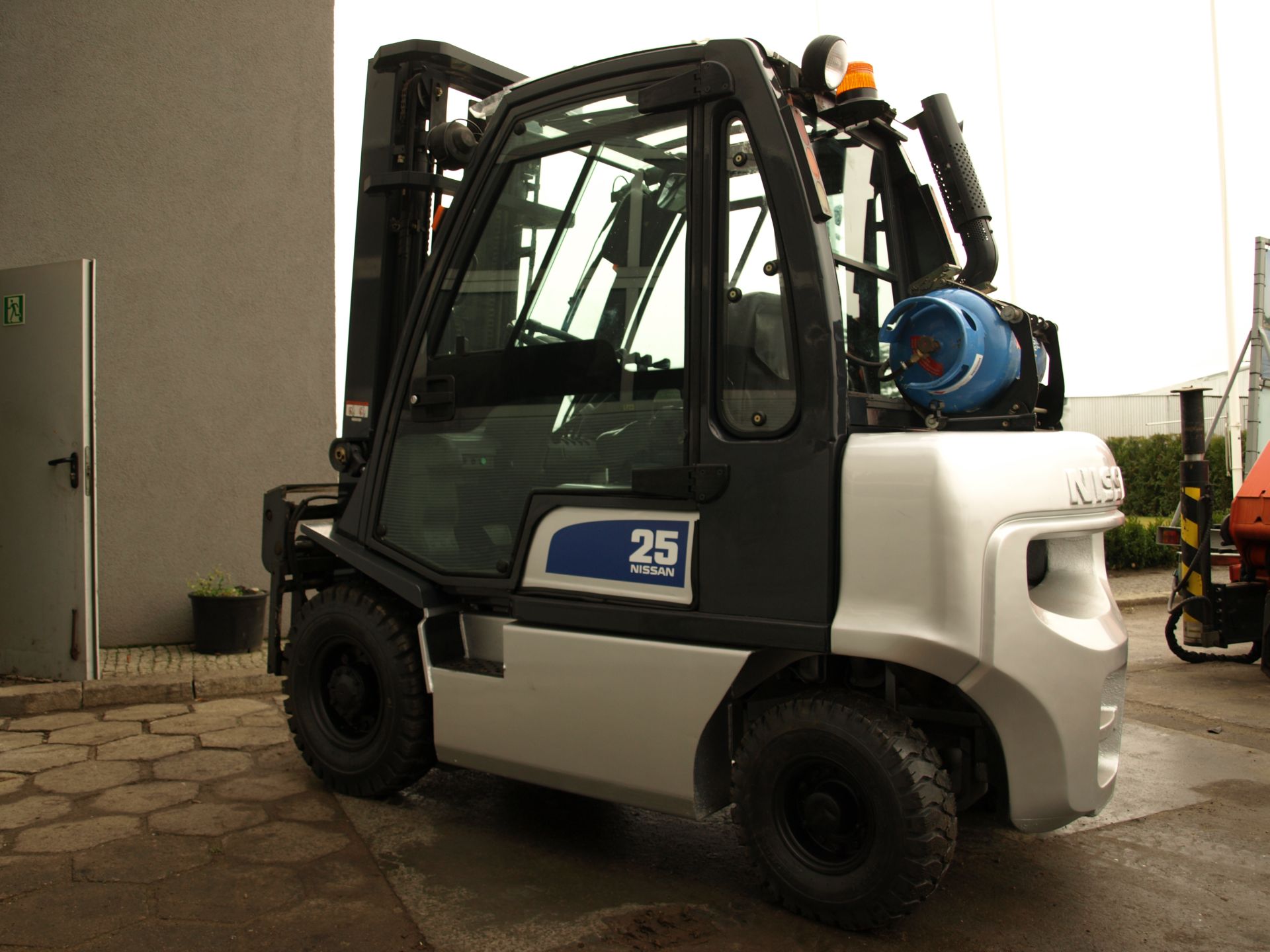 Nowy wózek widłowy spalinowy Nissan UD02A25PQ, srebrno-czarny, z niebieską butlą gazową, stojący na betonowym podłożu przy ścianie budynku.