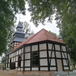 Kościół szachulcowy z czerwoną dachówką, widok z dołu przez korony drzew, z widoczną wieżą i krzyżem na szczycie, częściowo w trakcie renowacji.