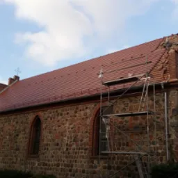 Murarz na rusztowaniu układa dachówkę na zabytkowym kościele z kamienia i cegły, widoczne krzyże na szczycie, częściowo wymienione poszycie dachu.