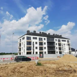 Nowoczesny, biały budynek wielorodzinny z ciemnymi ramami okiennymi i balkonami, widoczny z poziomu gruntu na placu budowy z piaskiem i ogrodzeniem tymczasowym.