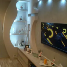 Nowoczesny salon z półkami w ścianie o nieregularnym kształcie, podświetleniem punktowym i telewizorem na ścianie, dekoracje na półkach i komodzie.