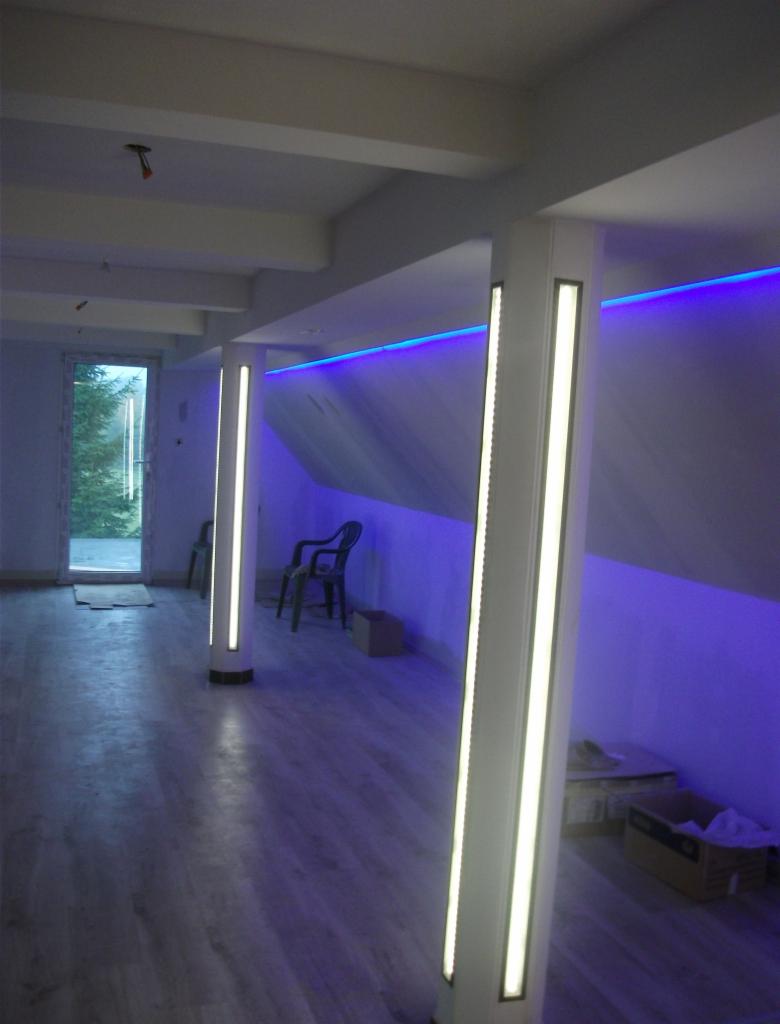 Wnętrze pokoju z panelami podłogowymi i kolumnami oświetleniowymi LED, niebieskie podświetlenie sufitu, widoczne kartony i krzesła w tle.