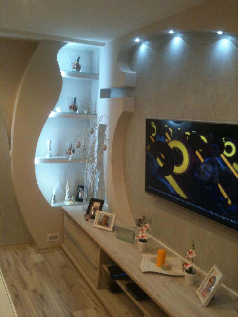 Nowoczesny salon z półkami w ścianie o nieregularnym kształcie, podświetleniem punktowym i telewizorem na ścianie, dekoracje na półkach i komodzie.