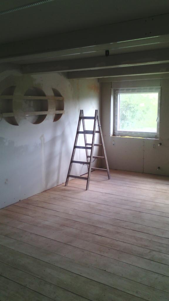 Wnętrze w trakcie prac: surowe drewniane deski podłogowe, ściany z częściowo położoną płytą kartonowo-gipsową, drabina oparta o ścianę, okno w tle, w ścianie wnęki w kształcie półksiężyców.