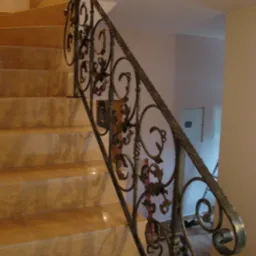 Kuta, ozdobna balustrada schodowa z metalu w kolorze ciemnego brązu, zamontowana przy schodach wyłożonych płytkami w odcieniach beżu.