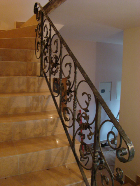 Kuta, ozdobna balustrada schodowa z metalu w kolorze ciemnego brązu, zamontowana przy schodach wyłożonych płytkami w odcieniach beżu.