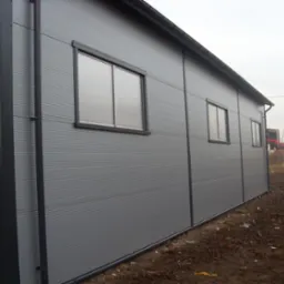 Szara hala z płyty warstwowej, widok z zewnątrz. Widoczne okna z ciemnymi ramami, rynna i fragment terenu.