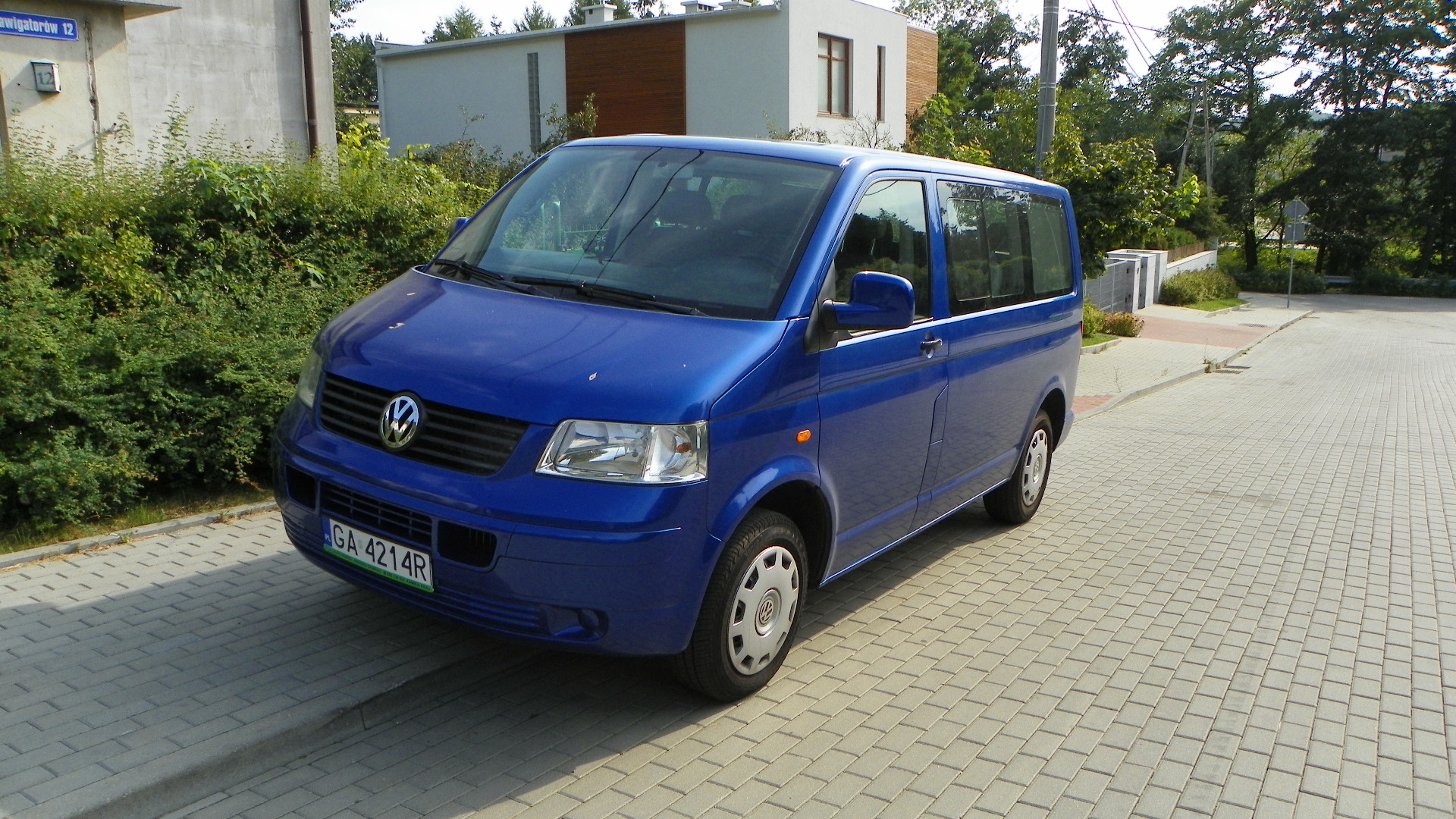 Niebieski Volkswagen Transporter T5 zaparkowany na chodniku z kostki brukowej, na tle nowoczesnego budynku i zieleni.