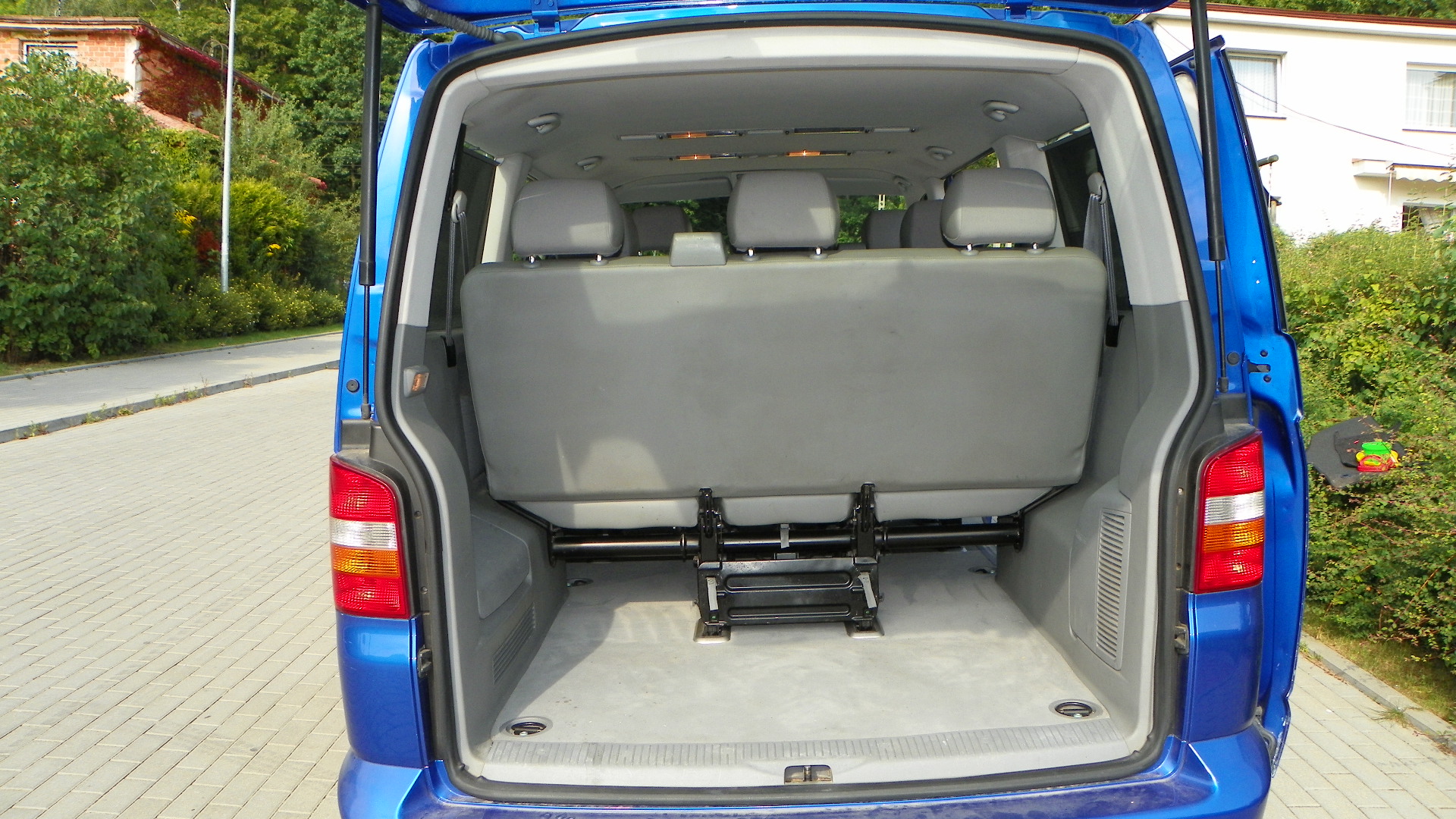 Wnętrze bagażnika niebieskiego busa z widocznymi szarymi fotelami i metalową konstrukcją mocującą, otwarty bagażnik na tle kostki brukowej i zieleni.