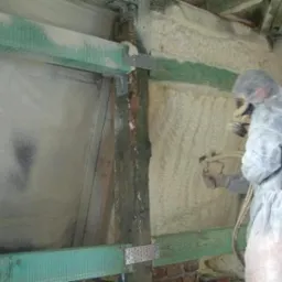 Pracownik w kombinezonie ochronnym natryskuje pianę izolacyjną na ścianę i sufit poddasza, widoczne drewniane elementy konstrukcyjne.