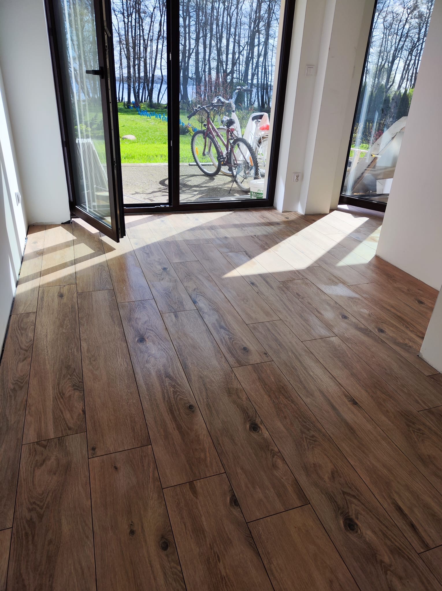 Ułożona, brązowa podłoga z paneli imitujących drewno, widok na otwarte okno balkonowe z rowerem na zewnątrz i promienie słoneczne na podłodze.