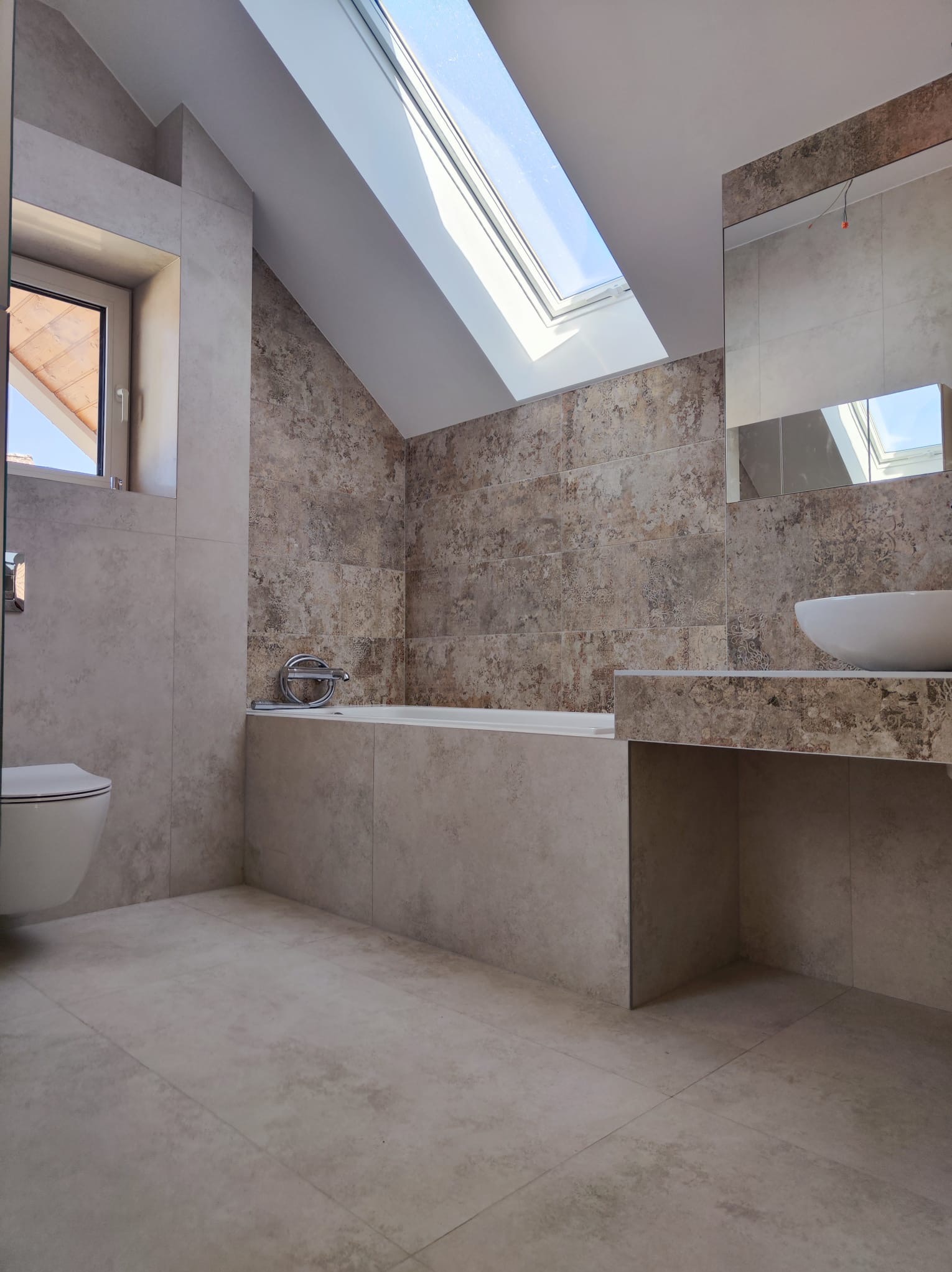 Jasna łazienka na poddaszu z oknem dachowym, beżowe płytki imitujące kamień na ścianach i podłodze, wanna zabudowana płytkami, umywalka nablatowa i toaleta.