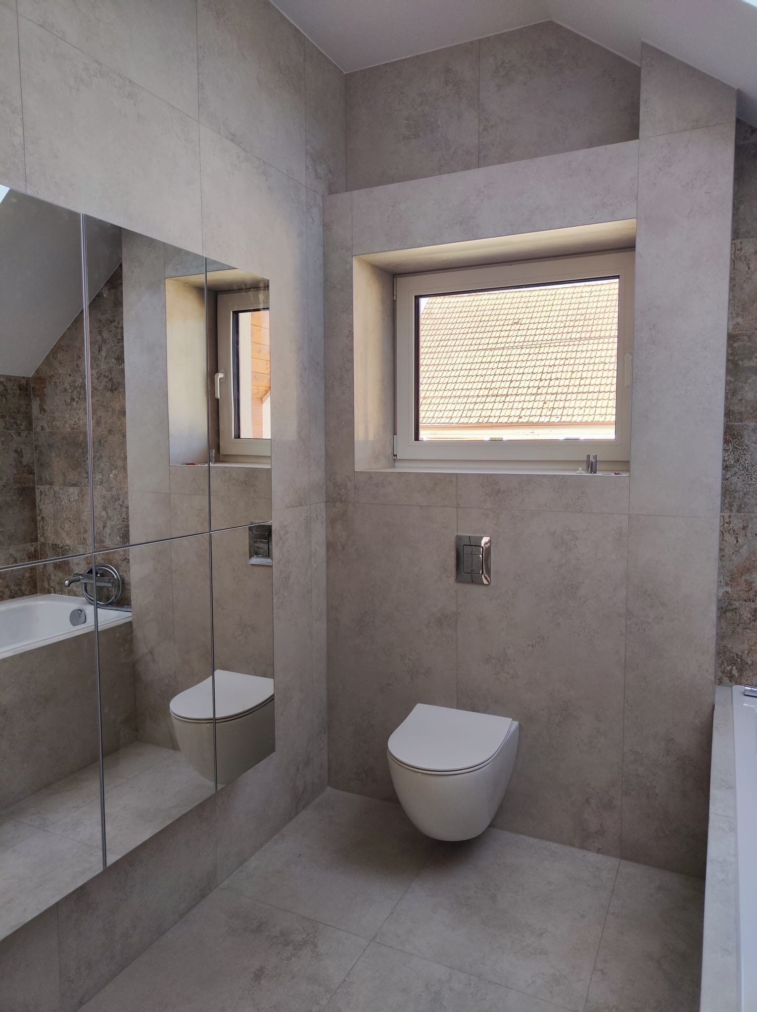 Nowoczesna łazienka z szarymi płytkami imitującymi beton, wisząca toaleta z chromowanym przyciskiem, duże lustro z odbiciem wanny i okno z widokiem na dach.