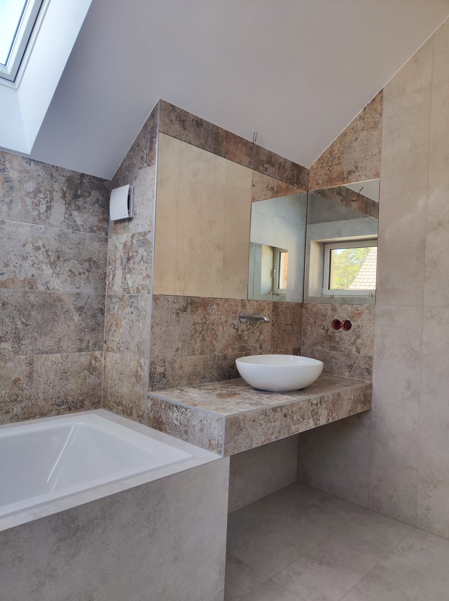 Nowoczesna łazienka z wanną, umywalką nablatową i lustrem, wykończona płytkami w odcieniach beżu i brązu z delikatnym wzorem, doświetlona oknem dachowym.