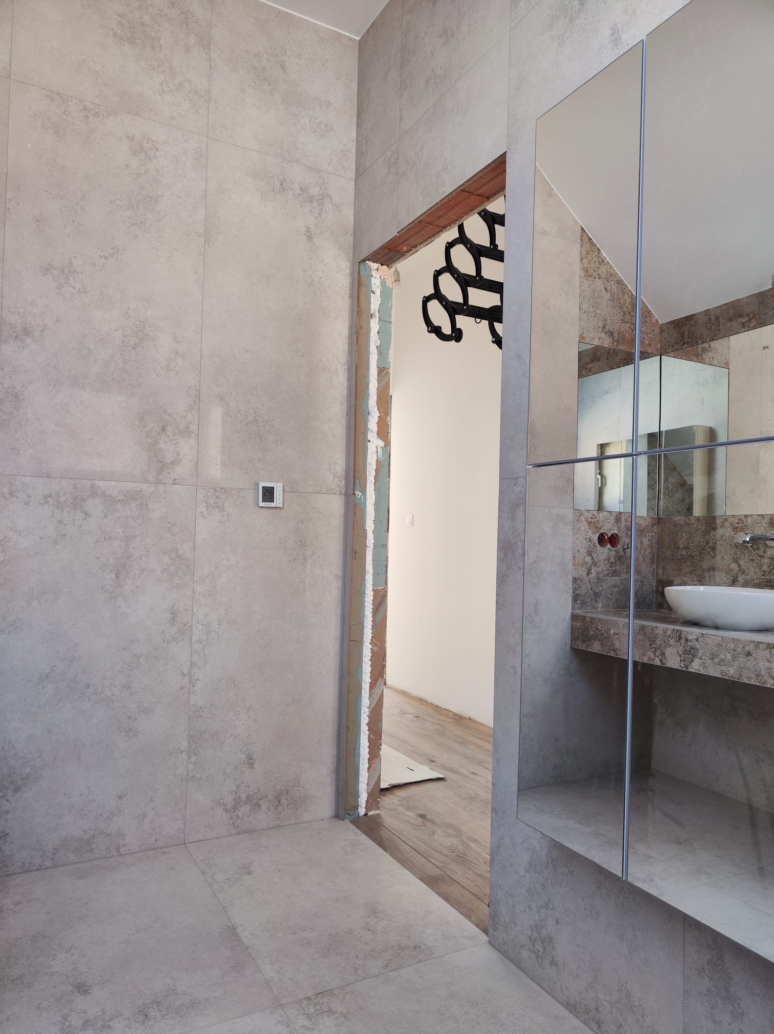 Łazienka w trakcie remontu: ściany i podłoga wyłożone szarymi płytkami imitującymi kamień, widoczny fragment montowanego lustra nad umywalką, surowy otwór drzwiowy z widoczną konstrukcją ościeżnicy...