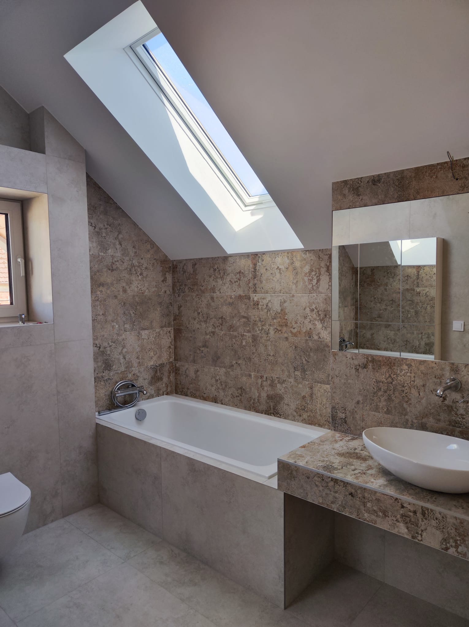 Nowoczesna łazienka na poddaszu z oknem dachowym, beżowe płytki imitujące kamień, biała wanna i umywalka nablatowa, lustro z szafką, minimalistyczny styl.