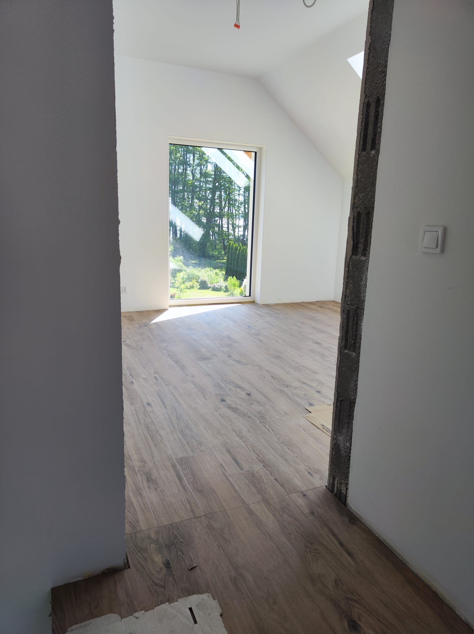Układanie podłogi z paneli imitujących drewno w jasnym pokoju z oknem na ogród, widoczny surowy otwór drzwiowy i fragment pomalowanej ściany.