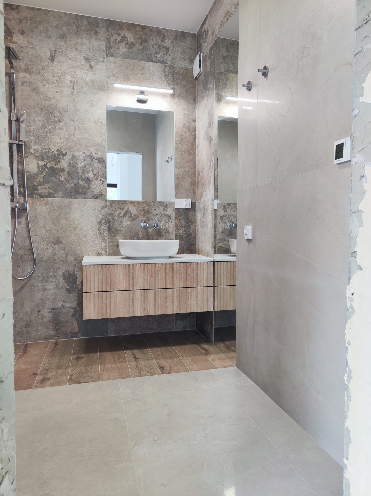 Nowoczesna łazienka z umywalką nablatową, drewnianą szafką z szufladami i mieszanką płytek w odcieniach szarości i beżu, widoczne fragmenty niedokończonego wykończenia przy ścianie.