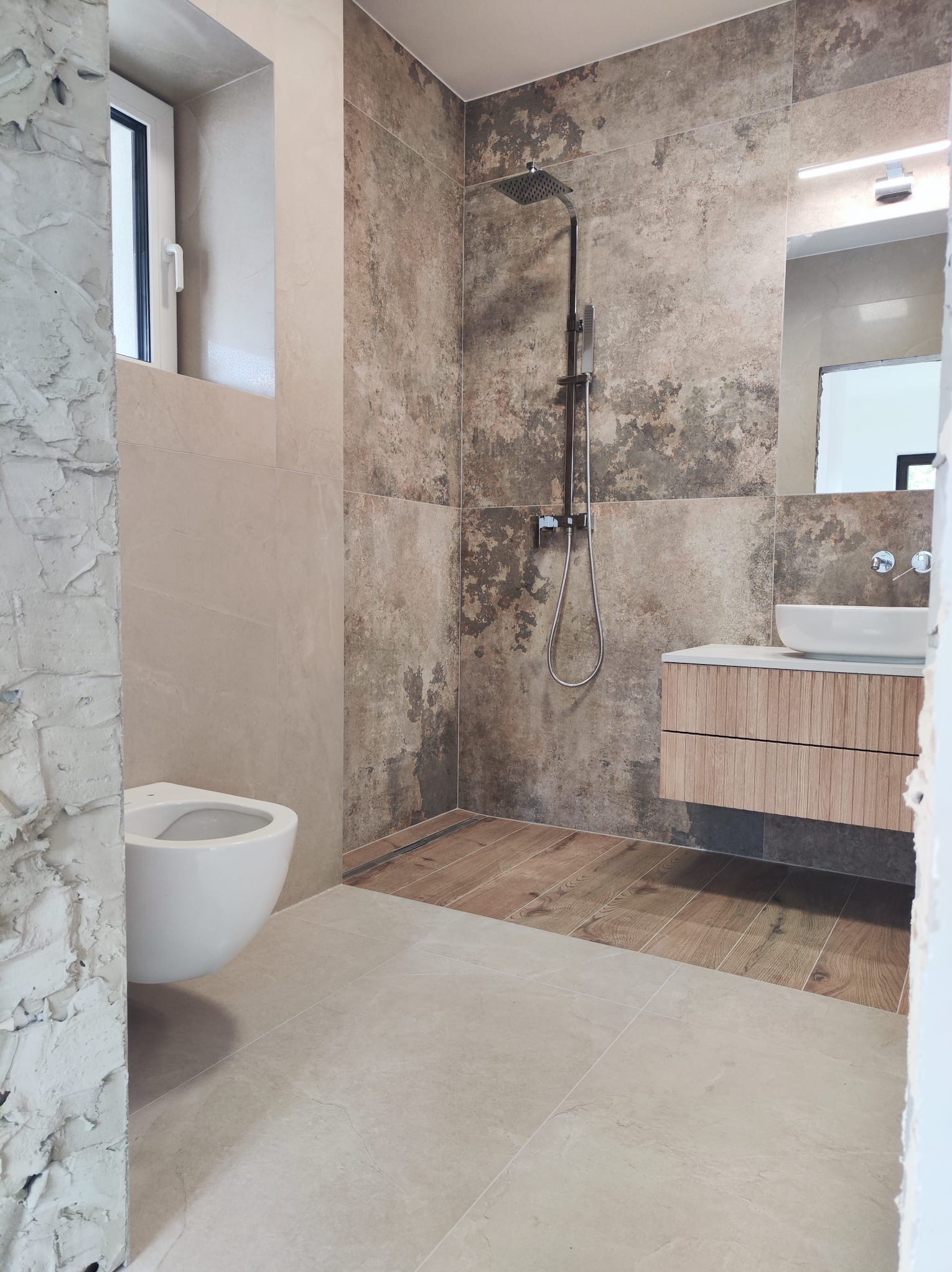 Nowoczesna łazienka w Dobrzanach w trakcie wykańczania: widoczna toaleta podwieszana, kabina prysznicowa typu walk-in z deszczownicą oraz szafka z umywalką nablatową, na ścianach płytki imitujące...