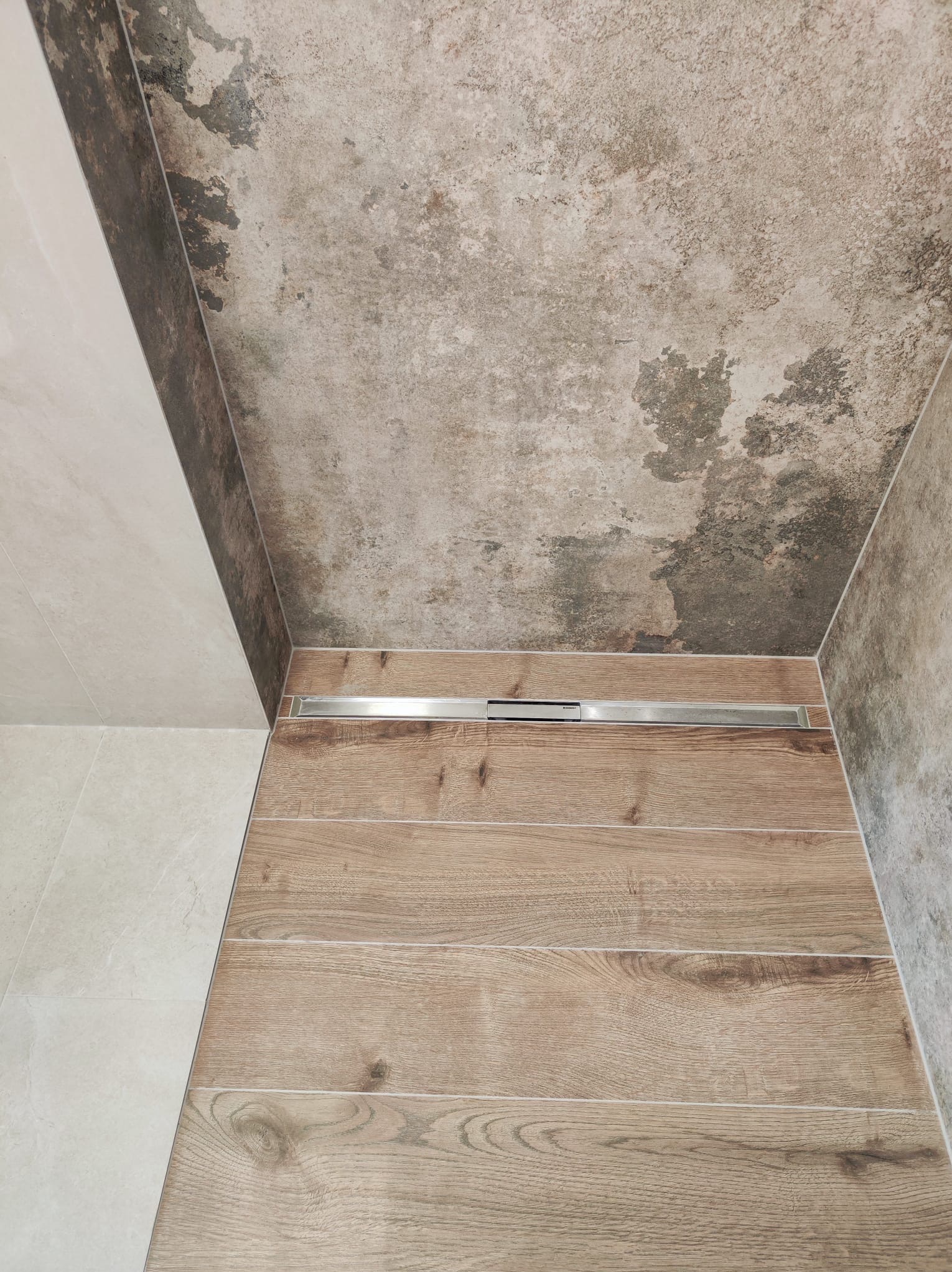 Nowoczesna kabina prysznicowa z odpływem liniowym, wykończona drewnopodobnymi płytkami na podłodze i szarą, betonową fakturą na ścianach, kontrastująca z jasnymi płytkami po lewej stronie.