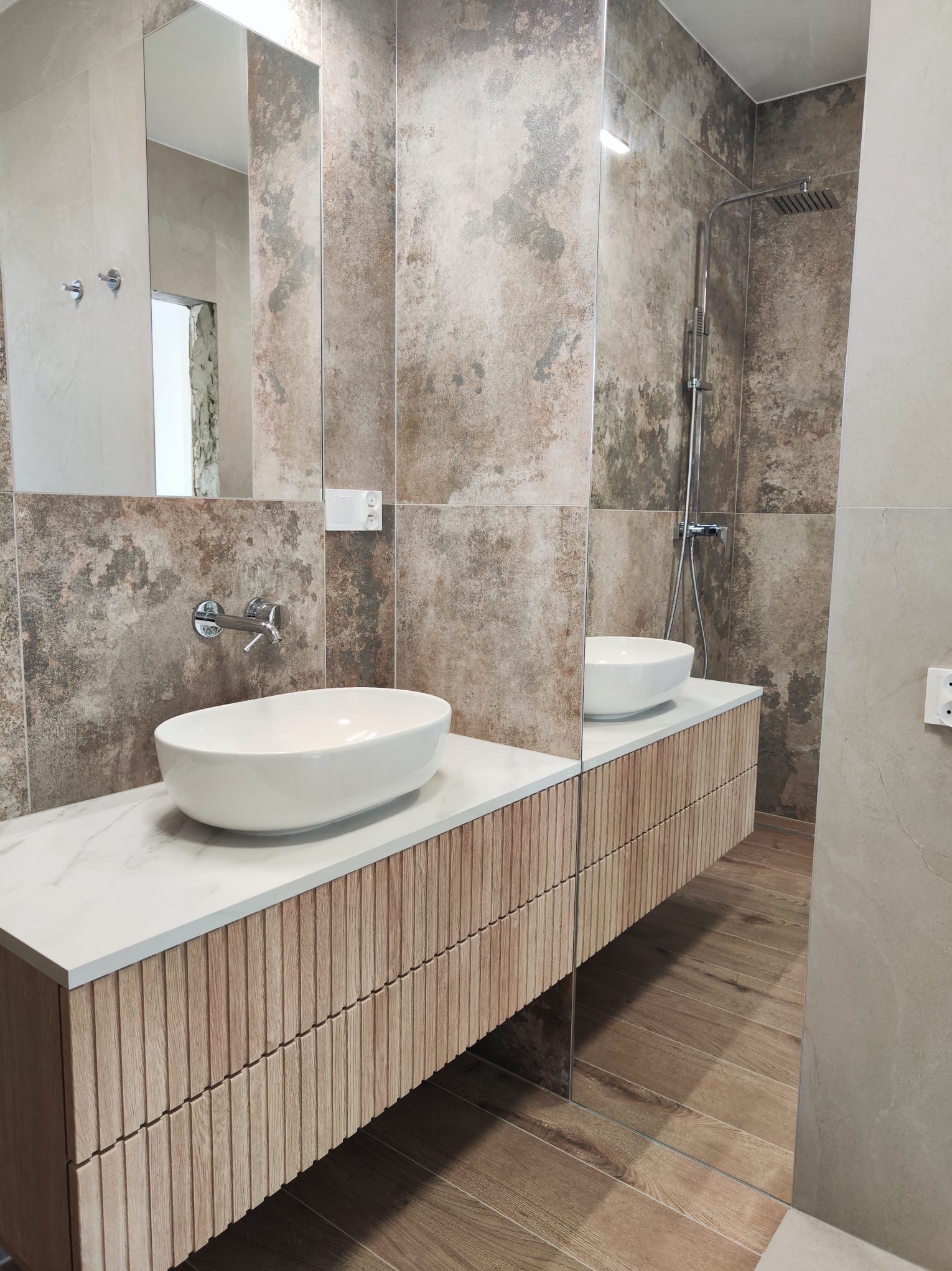 Nowoczesna łazienka z umywalką nablatową, drewnianą szafką z frezowanymi frontami, dużym lustrem bez ramy i płytkami imitującymi kamień na ścianach oraz drewnopodobnymi na podłodze.