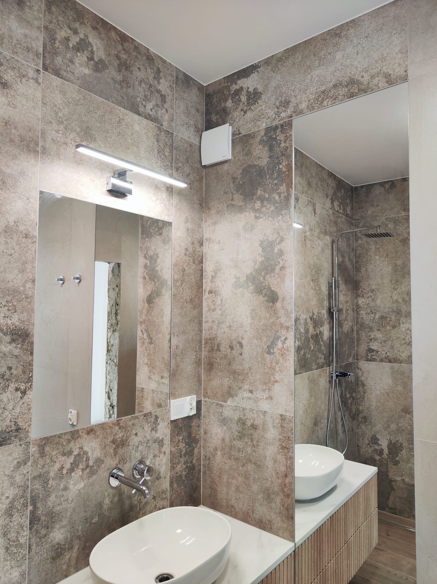 Nowoczesna łazienka z umywalkami nablatowymi, dużymi płytkami imitującymi kamień w odcieniach szarości i brązu, minimalistyczną armaturą i oświetleniem LED.