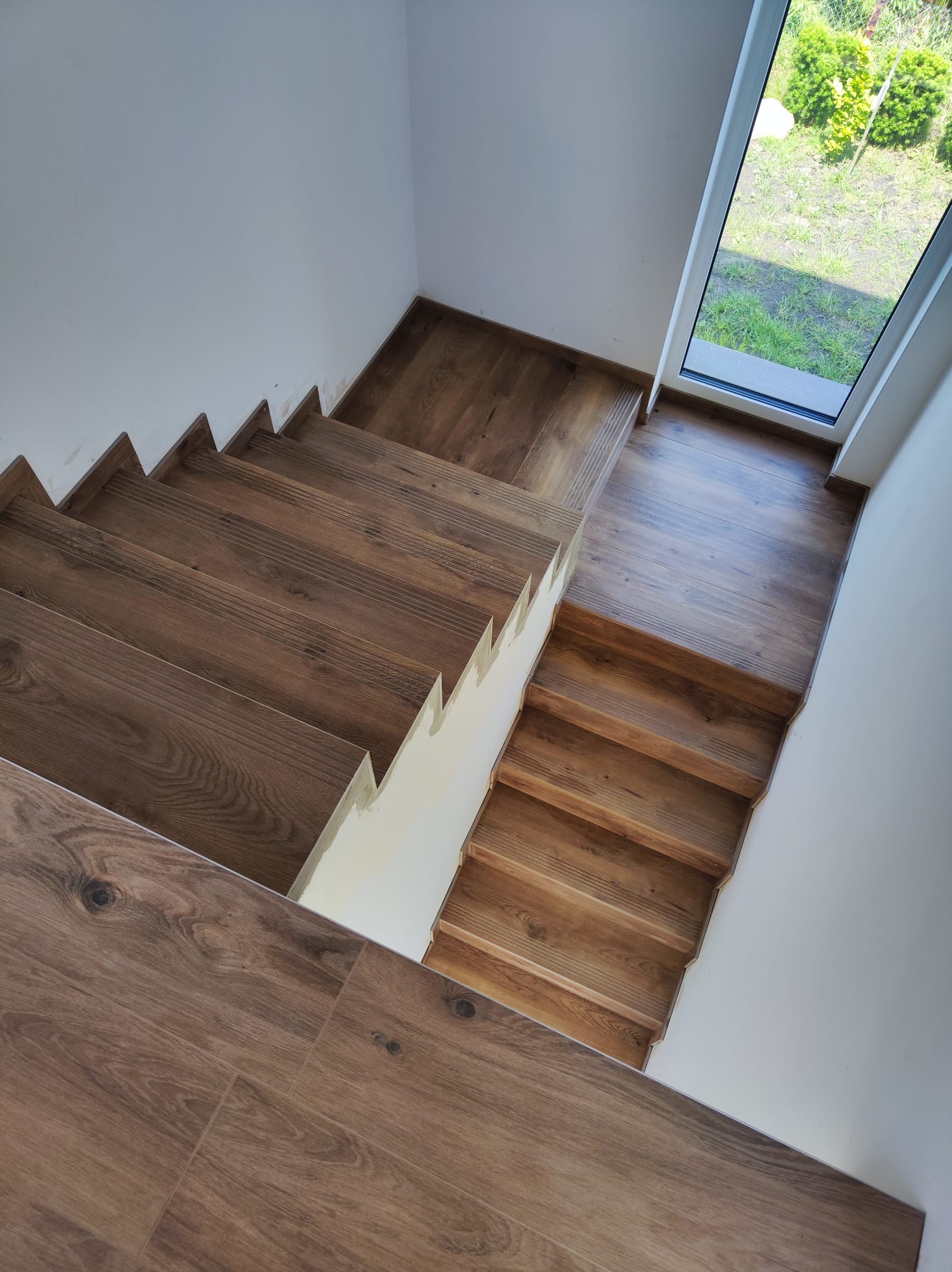 Klatka schodowa wyłożona panelami drewnopodobnymi, widok z góry, na ścianach widoczne ślady po montażu, okno z widokiem na zieleń.