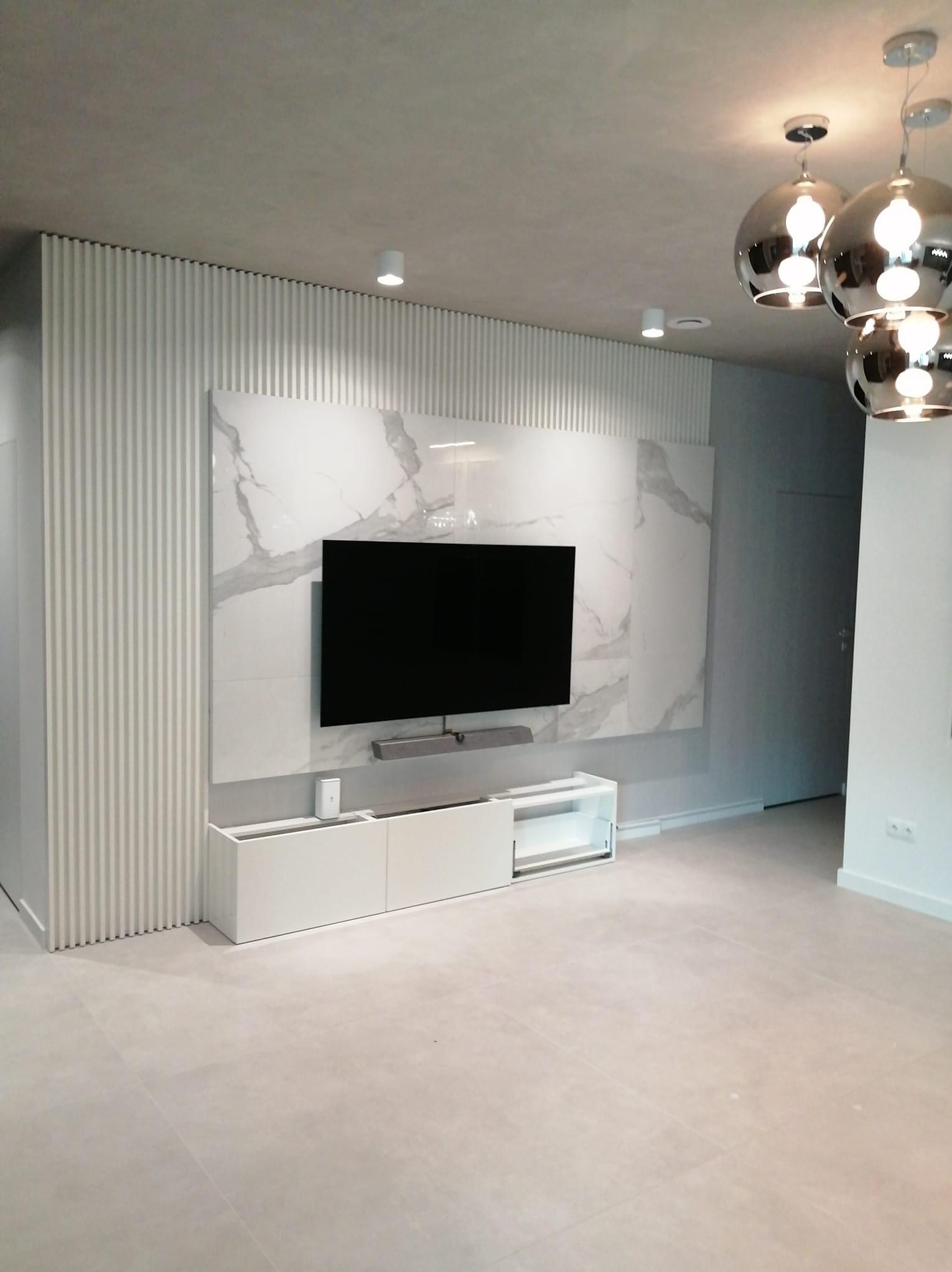 Nowoczesny salon z białą ścianą TV wykończoną płytami imitującymi marmur, dekoracyjnymi panelami ściennymi oraz wiszącymi lampami w stylu glamour.