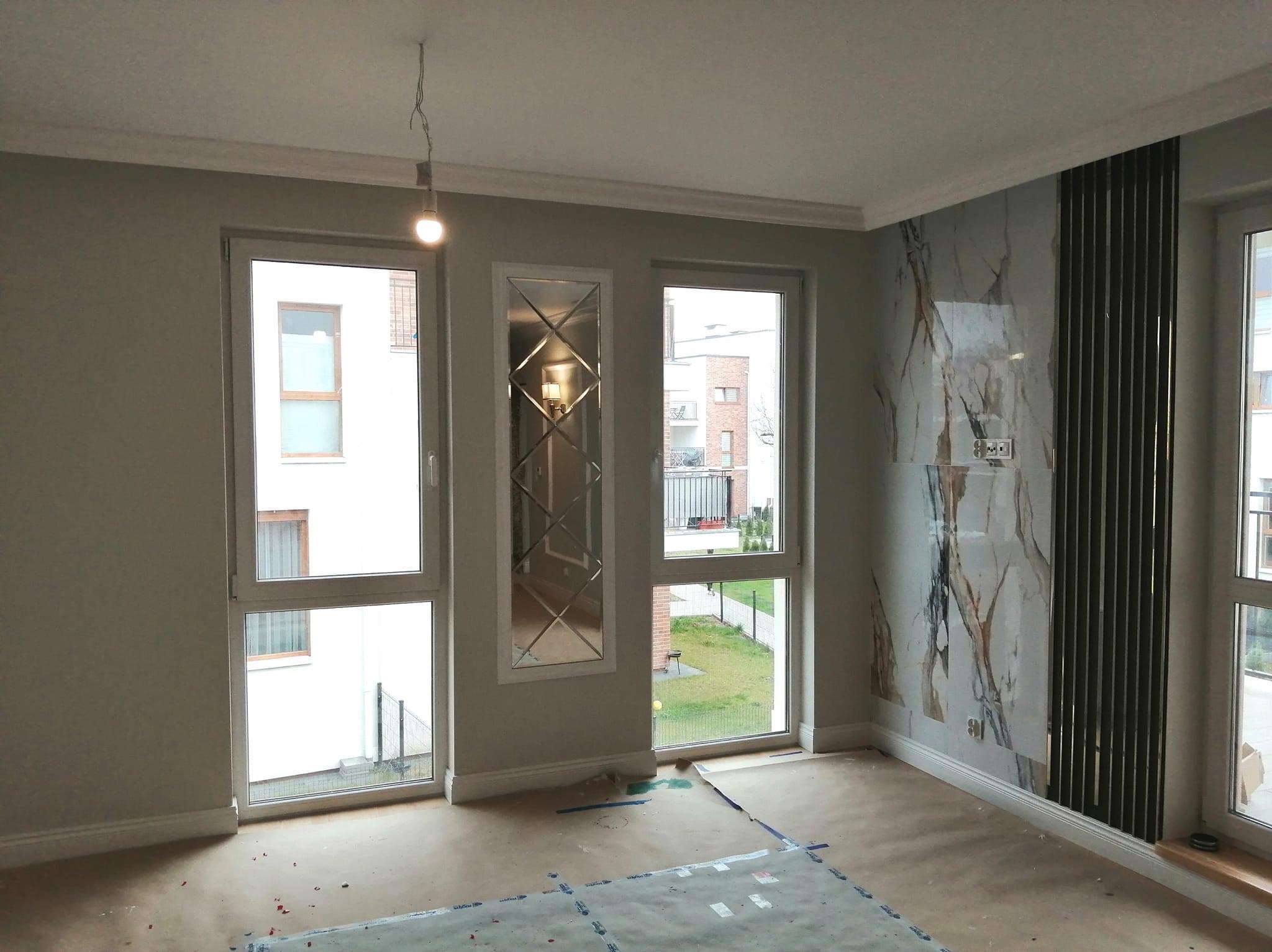 Pokój w trakcie prac wykończeniowych: zabezpieczona podłoga, ściany pomalowane na szaro, dekoracyjne lustro w ramie, marmurowa okładzina na ścianie z zamontowanym grzejnikiem panelowym, widoczne...