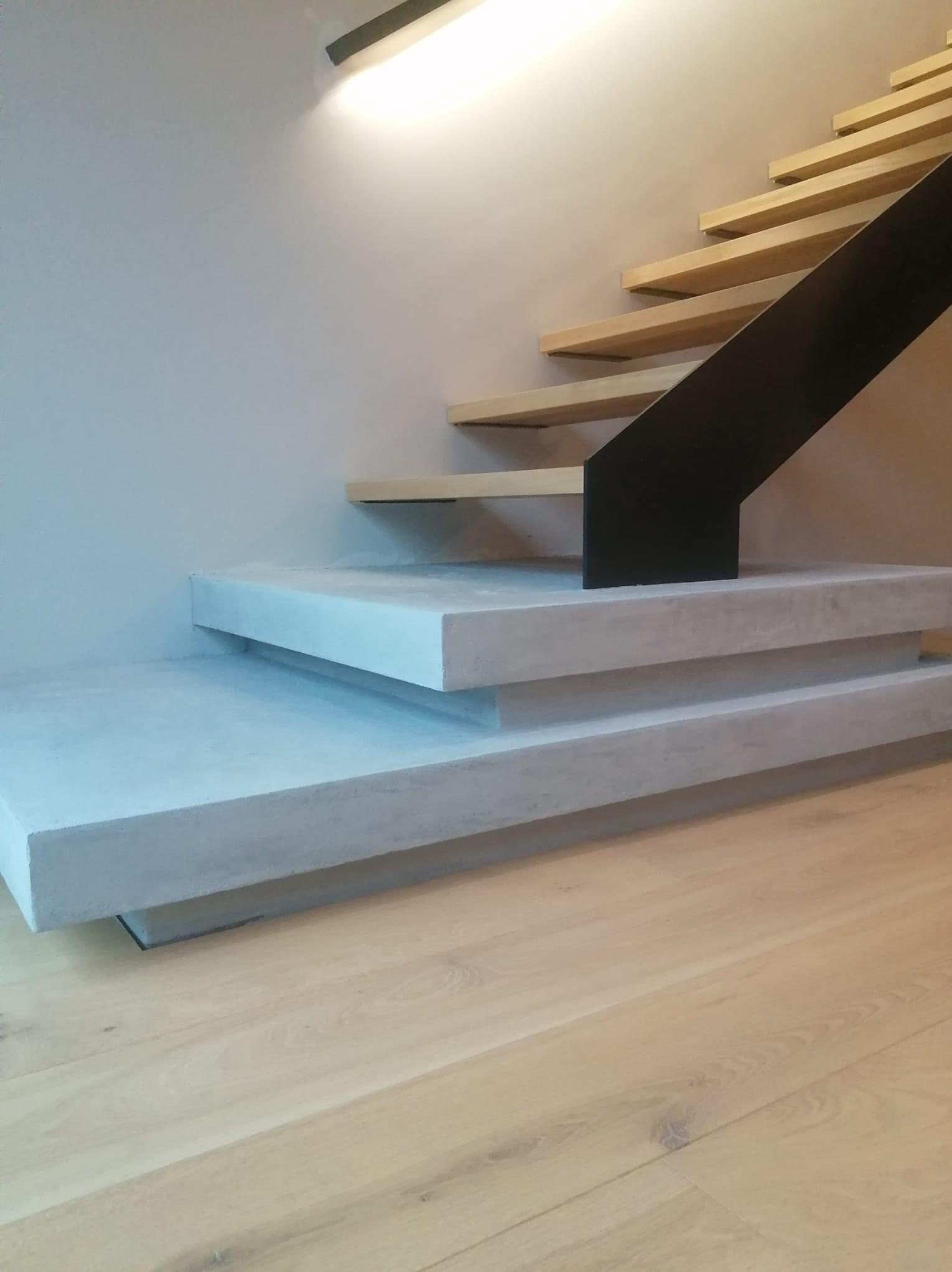 Nowoczesne schody z drewnianymi stopniami i betonowymi podestami, czarna metalowa barierka, oświetlenie punktowe na ścianie, jasna drewniana podłoga.
