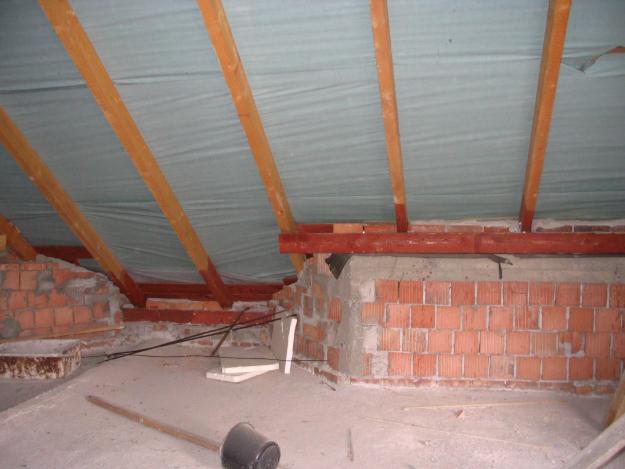 Surowe wnętrze poddasza w trakcie prac wykończeniowych: widoczne drewniane belki stropowe, izolacja, fragmenty murowanej ściany z czerwonej cegły oraz narzędzia budowlane, w tym wiadro i kij.
