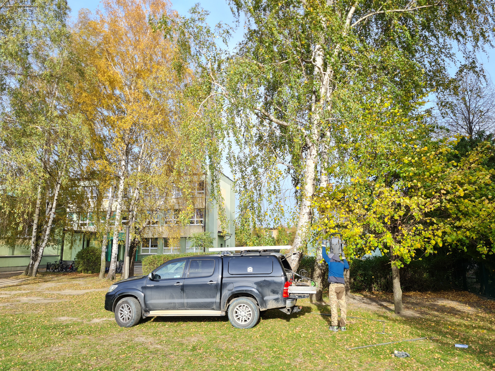 Pracownik w kamuflażu, oparty o brzozę, instaluje sprzęt pomiarowy, obok zaparkowany czarny pickup z zabudową i bagażnikiem dachowym.
