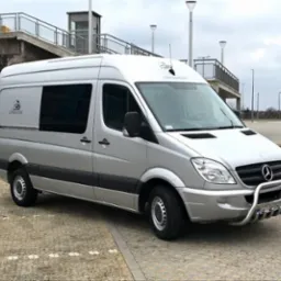 Srebrny Mercedes Sprinter z wysokim dachem, zaparkowany na kostce brukowej, z widocznym orurowaniem ochronnym z przodu i małym logo na boku, na tle wiaduktu i słupów oświetleniowych.