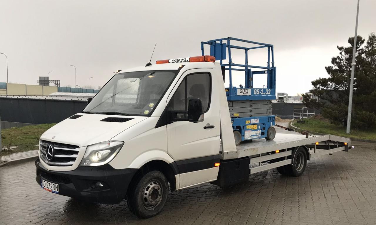 Biała laweta Mercedes-Benz z podnośnikiem nożycowym Gizo GS-1.32 na platformie, zaparkowana w Gdańsku na tle pochmurnego nieba i wiaduktu.