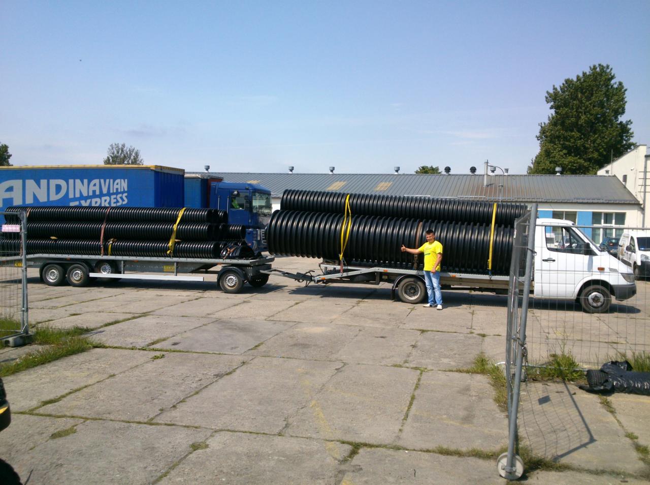 Dwa pojazdy, ciężarówka i bus z przyczepami, załadowane czarnymi rurami kanalizacyjnymi, zabezpieczonymi żółtymi pasami; mężczyzna w żółtej koszulce pokazuje kciuk w górę.