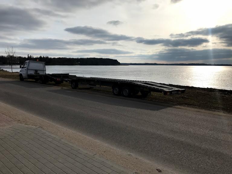 Biała ciężarówka z niskopodwoziową przyczepą stoi na poboczu asfaltowej drogi, z widokiem na jezioro i zachmurzone niebo w tle.