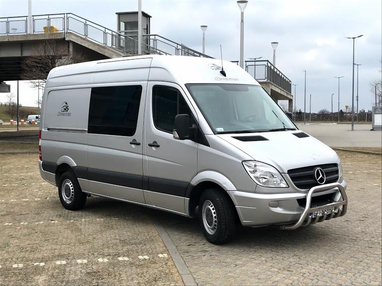 Srebrny Mercedes Sprinter z wysokim dachem, zaparkowany na kostce brukowej, z widocznym orurowaniem ochronnym z przodu i małym logo na boku, na tle wiaduktu i słupów oświetleniowych.