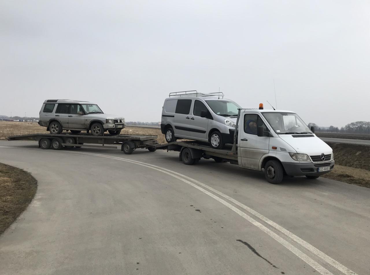 Laweta z dwoma pojazdami: srebrnym Land Rover Discovery na przyczepie i srebrnym Peugeot Expert na platformie holowniczej, sfotografowana na szarej drodze w pochmurny dzień.