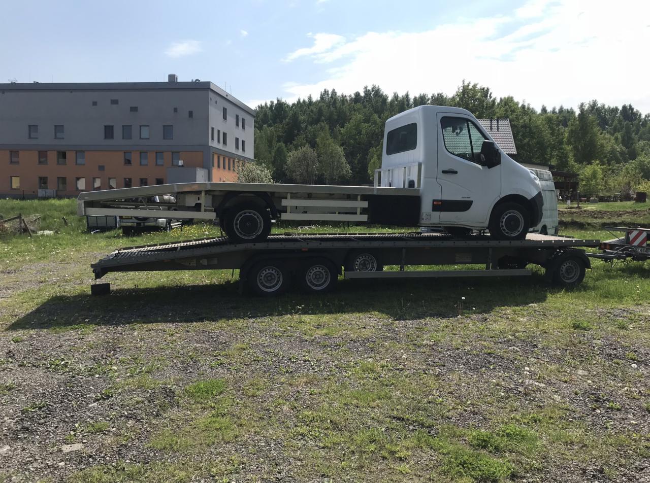 Biały samochód dostawczy na platformie lawety, zaparkowany na trawiastym terenie z budynkiem w tle.