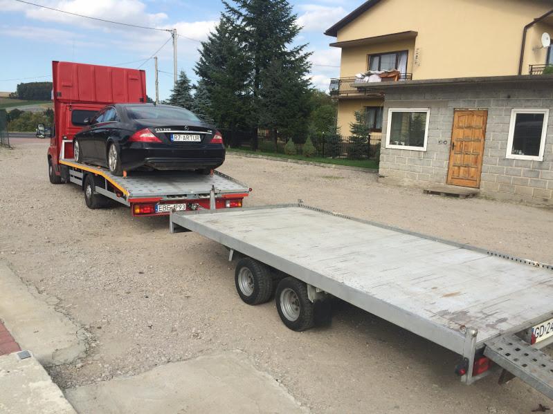 Czerwona ciężarówka z doczepioną lawetą transportuje czarny samochód osobowy Mercedes-Benz CLS 350 na tle zabudowań.