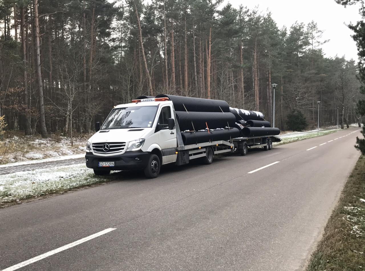 Biały Mercedes Sprinter z przyczepą przewożący duże czarne rury karbowane na tle leśnym, widoczne pomarańczowe światła ostrzegawcze na dachu.