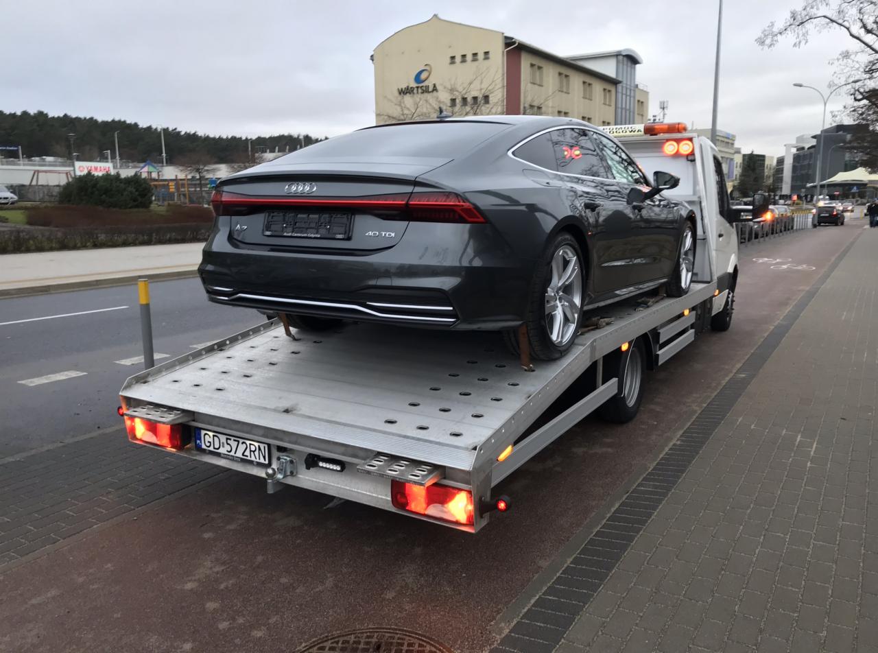 Szary samochód Audi A7 40 TDI, z widoczną tablicą rejestracyjną GD 572RN, umieszczony na platformie lawety na tle miejskiego krajobrazu z budynkiem firmy Wartsila.